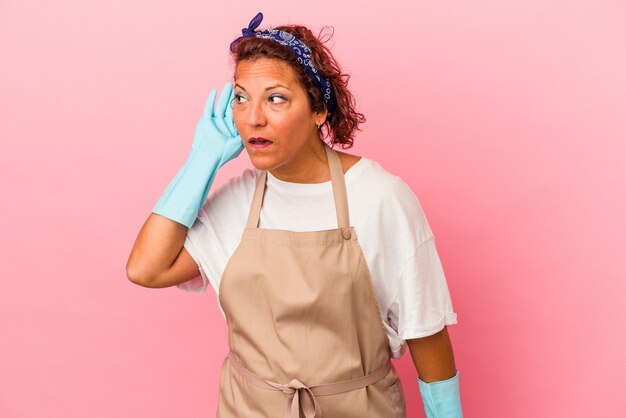 Latijnse vrouw van middelbare leeftijd geïsoleerd op roze achtergrond die een roddel probeert te luisteren.