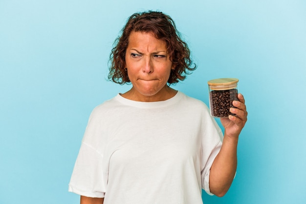 Latijnse vrouw van middelbare leeftijd met een koffiepot geïsoleerd op blauwe achtergrond verward, voelt zich twijfelachtig en onzeker.