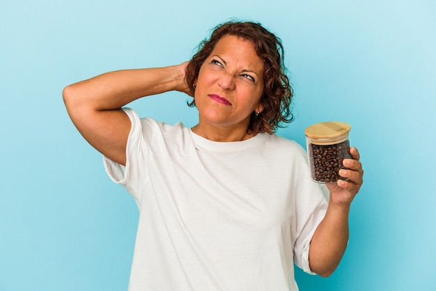 Latijnse vrouw van middelbare leeftijd met een koffiepot geïsoleerd op een blauwe achtergrond die de achterkant van het hoofd aanraakt, denkt en een keuze maakt.