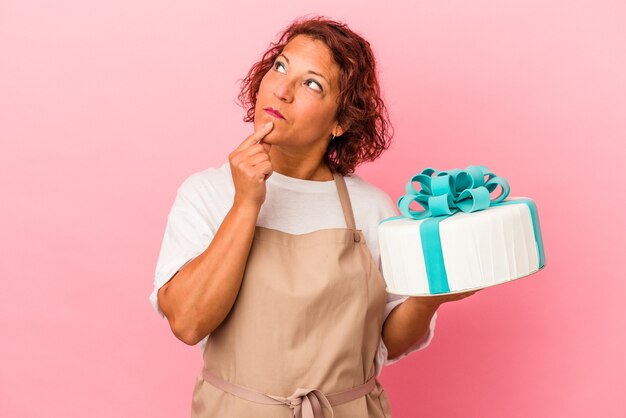 Latijnse vrouw van middelbare leeftijd met een taart die op een roze achtergrond wordt geïsoleerd en zijwaarts kijkt met een twijfelachtige en sceptische uitdrukking.