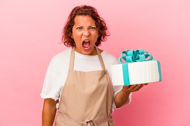 Latijnse vrouw van middelbare leeftijd met een taart geïsoleerd op roze achtergrond schreeuwend erg boos en agressief.