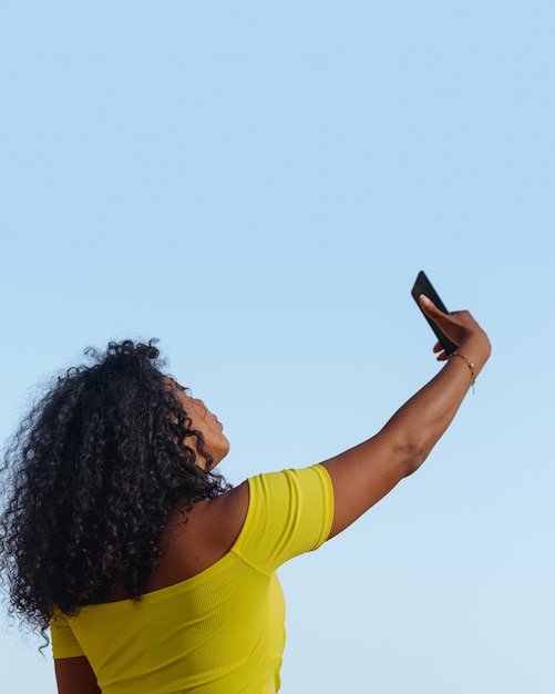 Latijnse zwarte vrouw maakt zelfportret met mobiel buitenshuis, blauwe lucht.