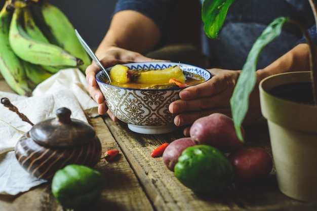 Latin American vegetable soup, hands grasping the plate. Ingredients carrot, chayote, sweet potato, green banana pepper.