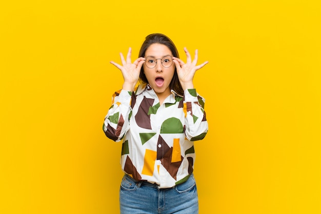 Latin american woman feeling shocked, amazed and surprised, holding glasses with astonished, disbelieving look isolated against yellow wall