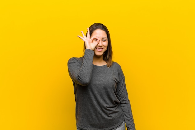 Latin american woman smiling happily with funny face, joking and looking through peephole, spying on secrets isolated against yellow wall