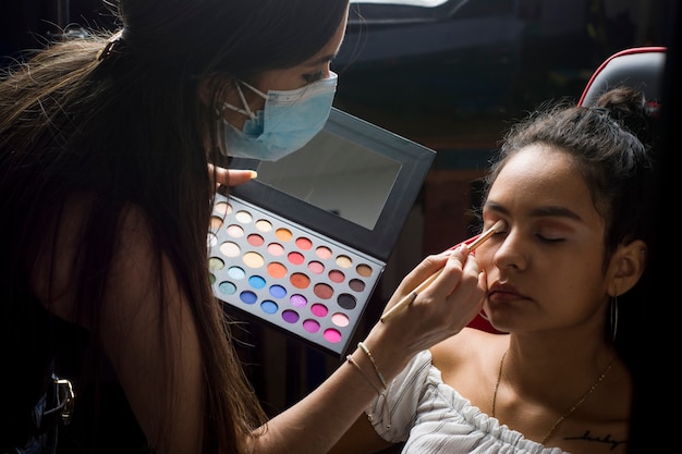 Молодая женщина из Латинской Америки наносит макияж на модель с протоколом биобезопасности антисовидного вируса