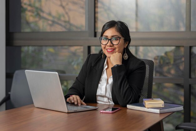 Латинская бизнес-женщина смотрит на камеру во время работы в офисе на своем ноутбуке с копировальным пространством