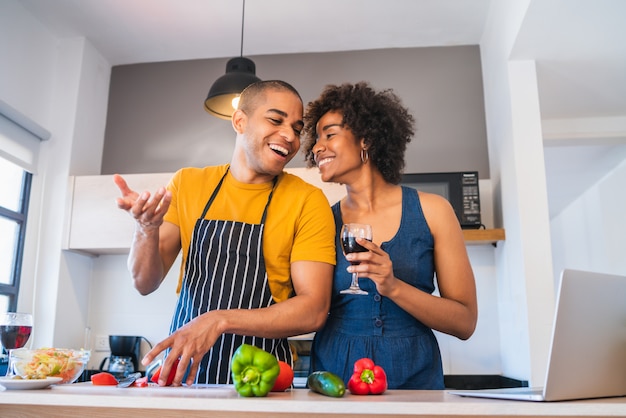 Latin couple using laptop while cooking in kitchen.