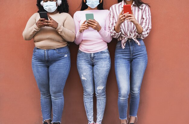 Ragazze latine che usano il cellulare in città mentre indossano una maschera di sicurezza per l'epidemia di coronavirus - giovani donne multirazziali con corpi e colori della pelle diversi