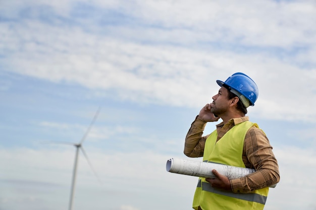 Latin male engineer standing on wind turbine field and talking on the phone