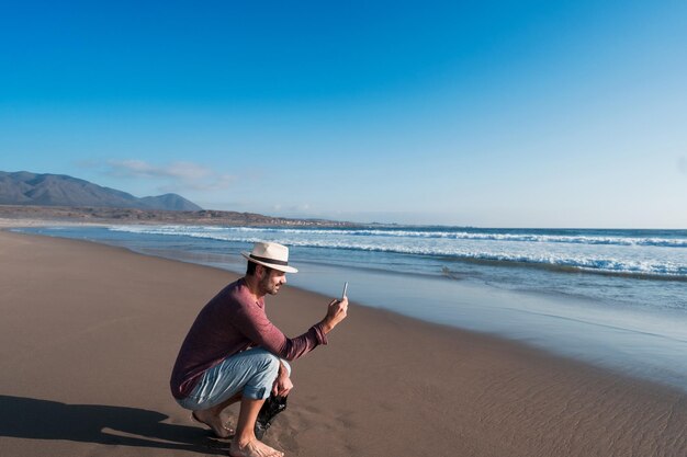 Uomo latino da solo sulla spiaggia che scatta foto con il suo cellulare al tramonto