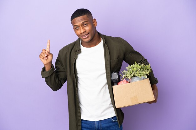 Photo latin man making a move while picking up a box full of things showing and lifting a finger in sign of the best