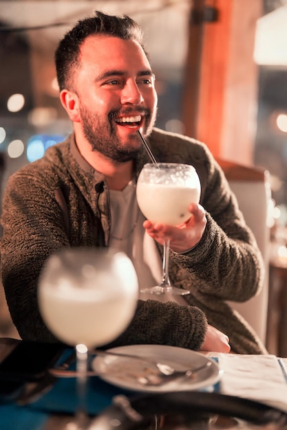 Latin man sitting in a bar or restaurant happily smiling and\
drinking