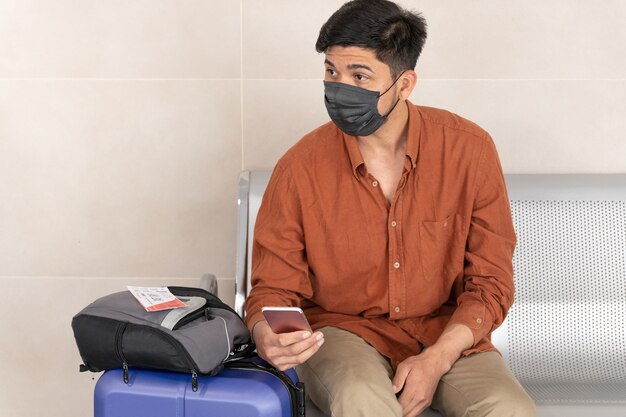 Latin man sitting at station with suitcase and using mobile phone