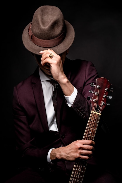 Latin man in suit with hat and guitar in hand drinking glass of wine sitting and looking at camera