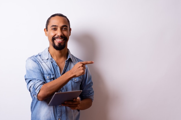 Latin man with great attitude points the finger at the empty space and holds a tablet in his hand on white background. Copy Space.