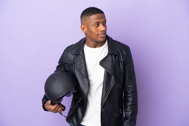 Latin man with a motorcycle helmet isolated on purple wall looking to the side and smiling