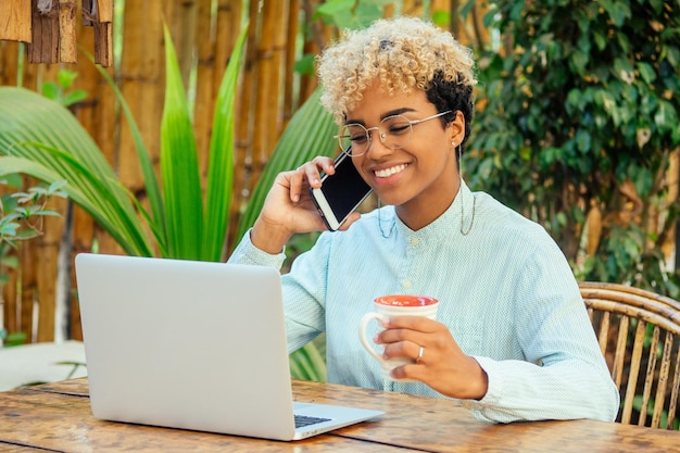 Latin mixed race curly woman looking at laptop and smiling at tropical beach cafe with tea cup She buying airline tickets online