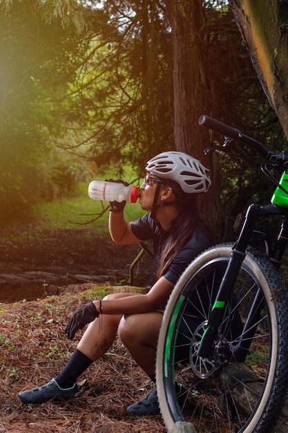 Latin woman cyclist in the middle of the forest sitting on a log hydrating while her bike is leaning against a tree