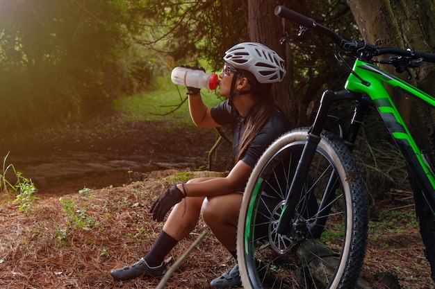 木にもたれて彼女の自転車で森の中で飲料水に座っているラテン系の女性サイクリスト