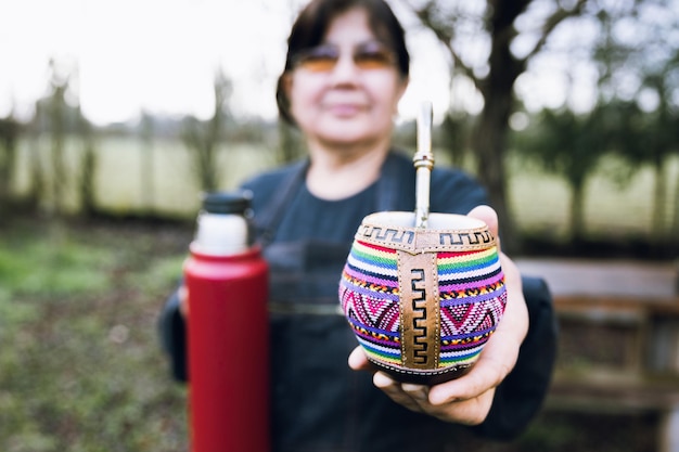 Latin woman serving mate with a red thermo, in a colorful ethnic mate.