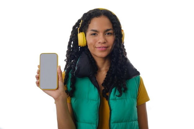 사진 고립 된 배경 위에 서있는 동안 카메라에 빈 화면으로 휴대 전화를 보여주는 라틴 여자. 광고 및 판촉 개념입니다.