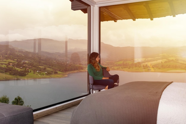 Latin woman sitting inside a room with large windows and a beautiful view of a lake and mountains at sunset