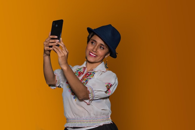 Latin woman in traditional dress and hat taking selfie picture with her cell phone while smiling in yellow background