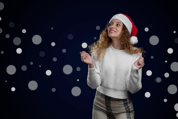 Latin woman wearing christmas hat and clothes pointing to a space for text with out of focus lights