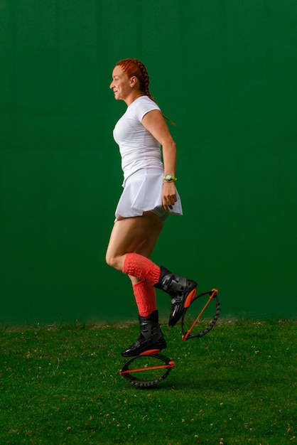 Latin woman, in white sportswear, doing an exercise routine wearing black Kangoo Jumps boots on dark green background