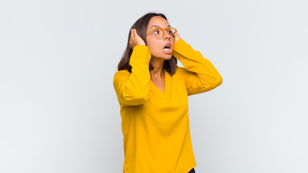 Latin woman with open mouth, looking horrified and shocked because of a terrible mistake, raising hands to head