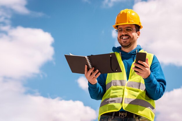 Latin worker with yellow hard hat, glasses and green vest, reading a book, a phone and smiling.