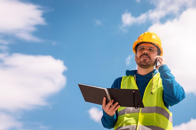 Latin worker with yellow hard hat, glasses and green vest, with a diary in his hands, talking on the phone and smiling.