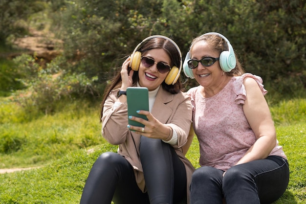 A Latin young adult woman and her mother sitting on the grass are listening to music