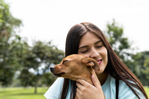 A Latin young adult woman hugs her female pinsher in the park