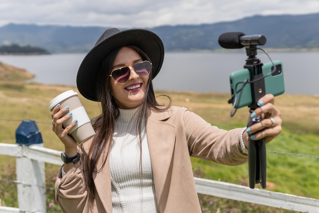 Photo a latin young adult woman with glasses coat and hat is streaming with her smartphone and promoting a coffee cup near at lake