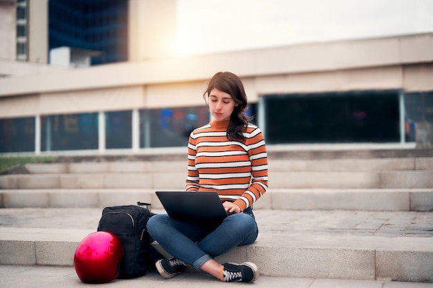 Latin young woman sitting outdoors on the stairs with a laptop and a bag