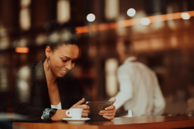 Latina woman in a coffee shop viewed through glass with reflections as they sit at a table chatting and laughing working on tablet computer