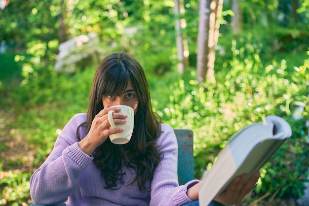 Latina giovane donna che legge un libro e beve da una tazza di caffè e guarda la telecamera