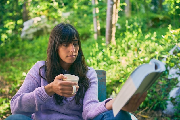 Latina young woman reading a book and holding a cup of coffee while sitting outdoors