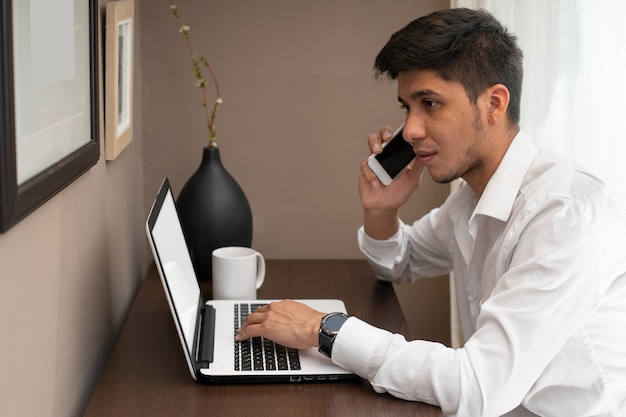 Latino man die op de mobiele telefoon praat terwijl hij thuis werkt vanaf een laptop