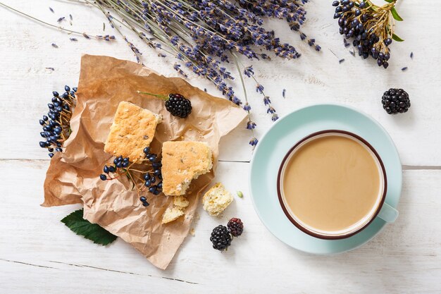 Latte, biscuits, berries and flowers composition. Blue coffee cup with creamy foam, dried flowers, cookies and blackberry at white wooden table, top view. Hot drinks, seasonal offer concept