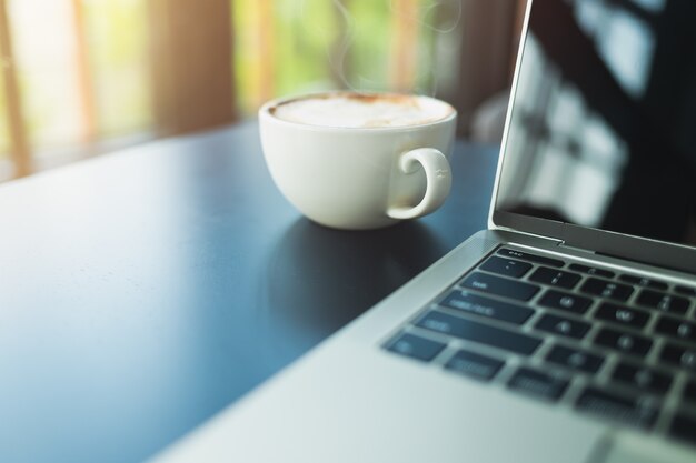 Кофе латте в белой кофейной кружке и на стороне ноутбука на черном деревянном столе