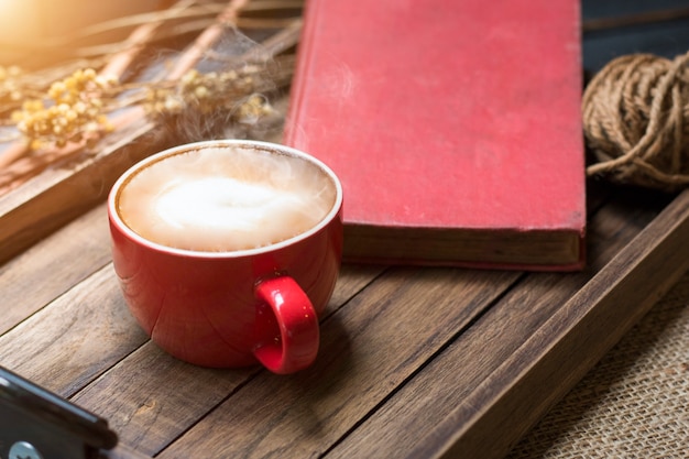 Чашка латте, книга на деревянном подносе с теплым утренним светом около окна.