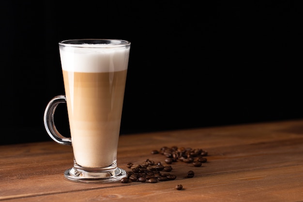 Latte macchiato in una tazza di vetro con manico con caffè intorno isolato su sfondo di legno