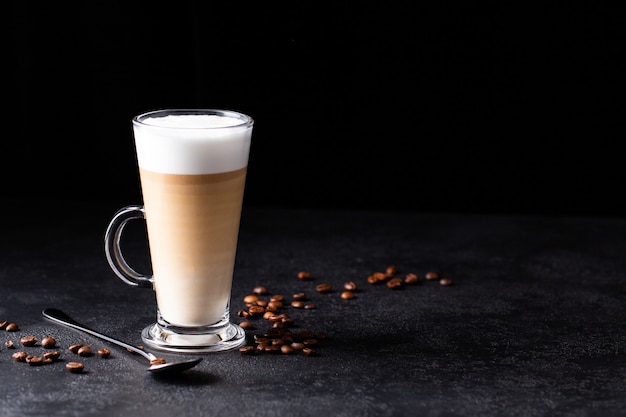 latte macchiato in a glass cup with handle with coffee around and spoon isolated on dark background