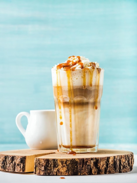 Latte macchiato with whipped cream and caramel sauce in tall glass on round wooden serving board over blue painted wall background