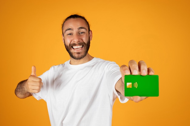 Laughing adult european man with beard in white tshirt hold credit card and thumb up