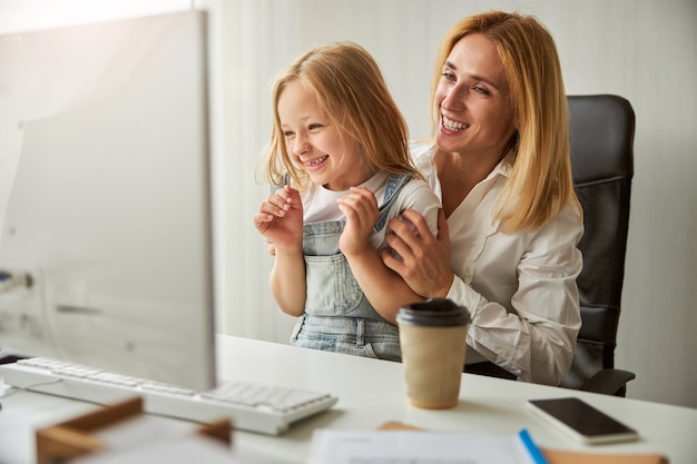 Laughing adult female with happy smiling daughter sitting at the work desk while looking at the screen of computer