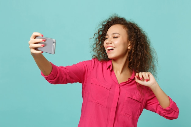 Laughing african girl in pink casual clothes doing selfie shot on mobile phone isolated on blue turquoise wall background in studio. People sincere emotions, lifestyle concept. Mock up copy space.